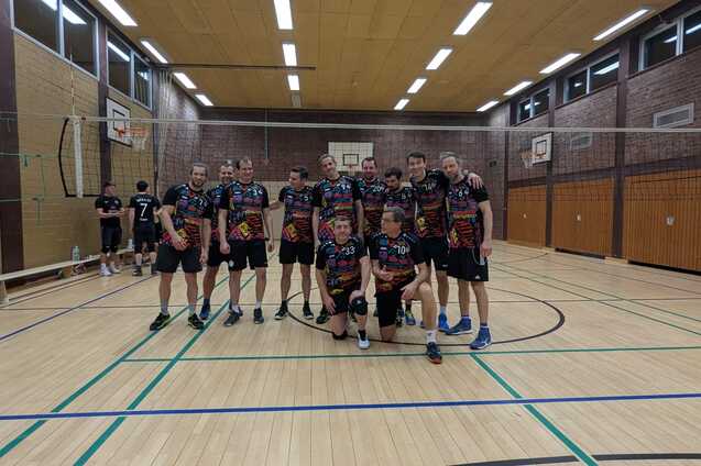 Gruppe von zehn Volleyballspielern in Sportkleidung posiert l&auml;chelnd in einer Halle mit Volleyballnetz im Hintergrund.