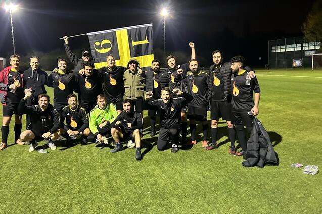 Feiernde Fu&szlig;ballmannschaft in schwarzen Trikots posiert mit einer Flagge auf dem Sportplatz bei Nacht.