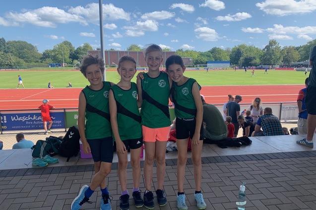 Vier Kinder in gr&uuml;nen Sporttrikots stehen l&auml;chelnd auf einer Trib&uuml;ne mit einer Laufbahn im Hintergrund.