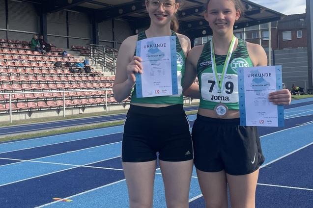 Zwei junge Athletinnen in Sportkleidung halten Urkunden und Medaille auf einer blauen Leichtathletikbahn w&auml;hrend eines Wettkampfs.