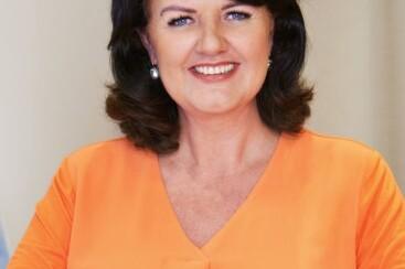 Portrait einer l&auml;chelnden Frau mit dunklen Haaren in einem orangefarbenen Oberteil, sitzend vor einem neutralen Hintergrund.