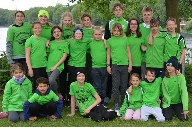 Gruppe von Jugendlichen in gr&uuml;nen T-Shirts steht am Ufer eines Gew&auml;ssers, umgeben von B&auml;umen.