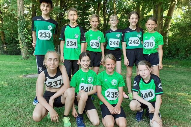 Gruppe junger Athleten in Sportkleidung mit Startnummern, auf einer Wiese im Freien, umgeben von B&auml;umen.