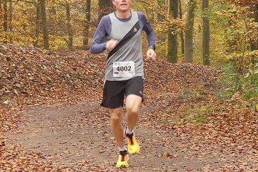 L&auml;ufer in Sportkleidung joggt auf einem mit Laub bedeckten Waldweg in einem herbstlichen Wald.