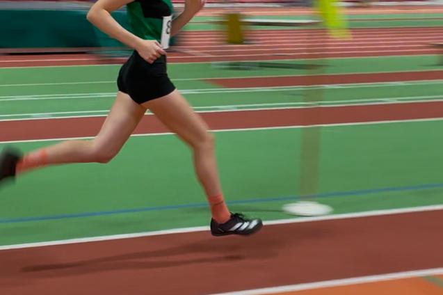 Junge Athletin sprintet auf einer leichtathletischen Bahn, im Hintergrund verschwommene Trib&uuml;nen und Hinweisschilder.