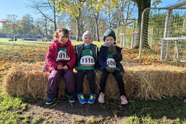 Drei Kinder in Sportkleidung mit Startnummern sitzen auf einem Strohballen in einem herbstlichen Garten.