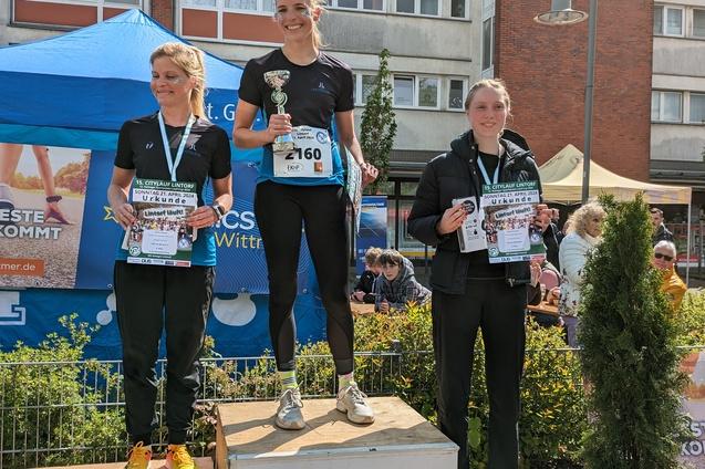 Drei Frauen stehen auf dem Siegertreppchen mit Pokal und Urkunden nach einem Wettlauf. Es ist eine sonnige Veranstaltung.