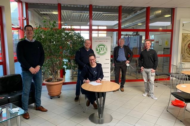 F&uuml;nf M&auml;nner stehen um einen Tisch, w&auml;hrend einer von ihnen etwas auf ein Dokument unterschreibt.