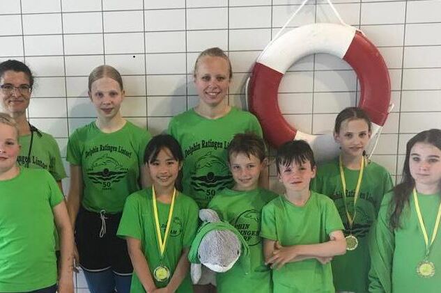Gruppe von acht Schwimmern in gr&uuml;nen T-Shirts, zwei halten Medaillen, vor einem Schwimmring an einer Wand.