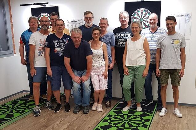 Gruppenfoto von 14 Personen im Darts-Raum mit zwei Dartboards und gr&uuml;nen Dartscheiben-Design-Matten.