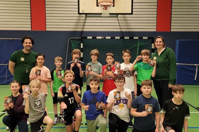 Gruppe von Kindern und Betreuern h&auml;lt Pokale in einer Sporthalle mit Basketballkorb im Hintergrund.