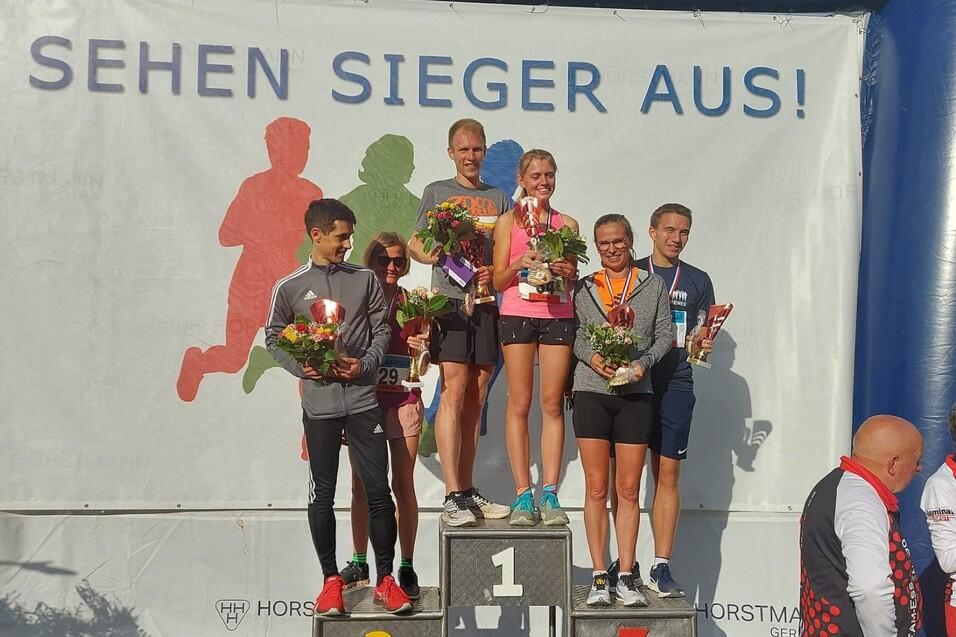 Siegerpodest mit sechs L&auml;ufern, die Medaillen und Blumen halten, vor einem Banner mit der Aufschrift &bdquo;SEHEN SIEGER AUS!&ldquo;.