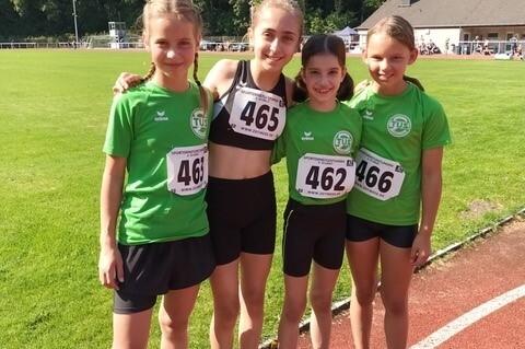 Vier junge Leichtathletinnen mit Startnummern auf einer Laufbahn in einem Stadion, gr&uuml;ne Vereins-T-Shirts und l&auml;chelnd.