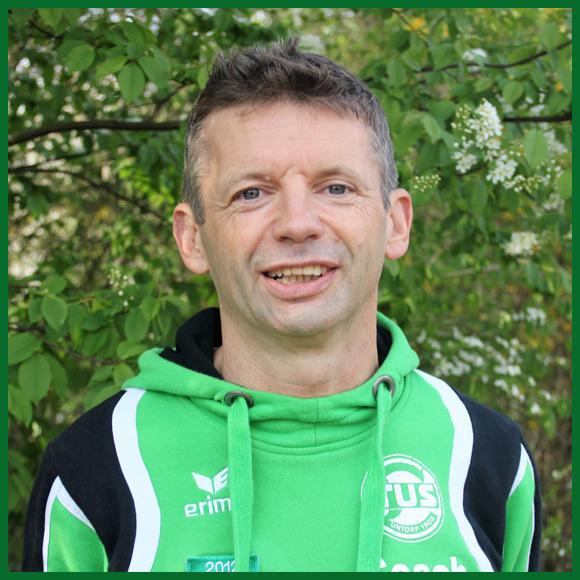 Portr&auml;t eines l&auml;chelnden Mannes in gr&uuml;ner Jacke vor einem Hintergrund aus Bl&auml;ttern und Bl&uuml;ten.