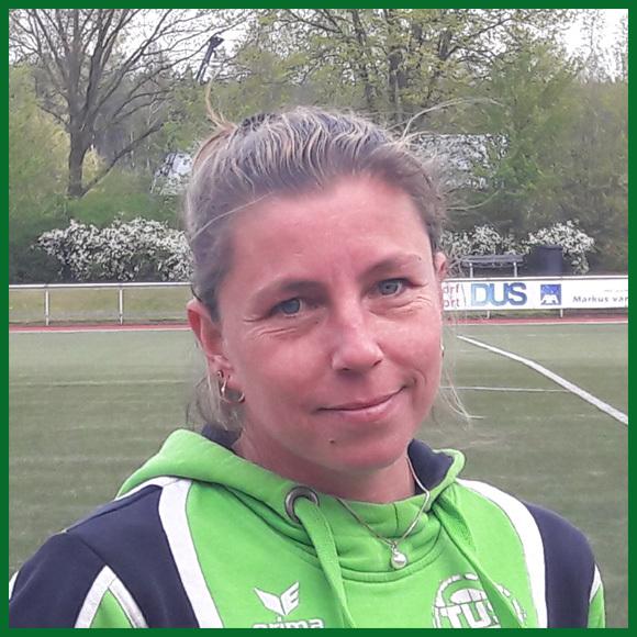 Portr&auml;t einer Frau mit kurzen, blonden Haaren in einem gr&uuml;nen Sweatshirt, Hintergrund mit Sportplatz und B&auml;umen.