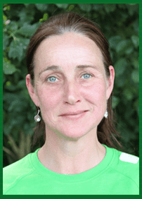 Portr&auml;t einer Frau mit braunen Haaren und blauen Augen, die ein gr&uuml;nes Shirt und Ohrringe tr&auml;gt, vor gr&uuml;ner Umgebung.