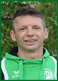 Portr&auml;t eines l&auml;chelnden Mannes in gr&uuml;ner Sportjacke vor gebl&uuml;mtem Hintergrund.