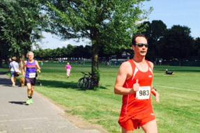 Läufer in rotem Outfit mit Startnummer 983 joggt auf einem Weg in einem Park, Bäume und Menschen im Hintergrund.