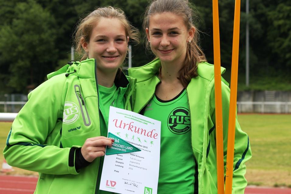 Zwei Jugendliche in gr&uuml;nen Sportjacken halten stolz eine Urkunde auf einem Sportplatz.