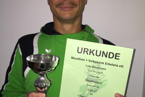 Mann in gr&uuml;ner Sportjacke h&auml;lt einen Pokal und eine Urkunde f&uuml;r den ersten Platz beim Marathonlauf.