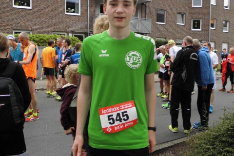 Junger Mann in gr&uuml;nem Sporttrikot mit Startnummer 545 steht in der Menge vor einem Wettkampf. Hintergrund mit L&auml;ufern und Geb&auml;uden.