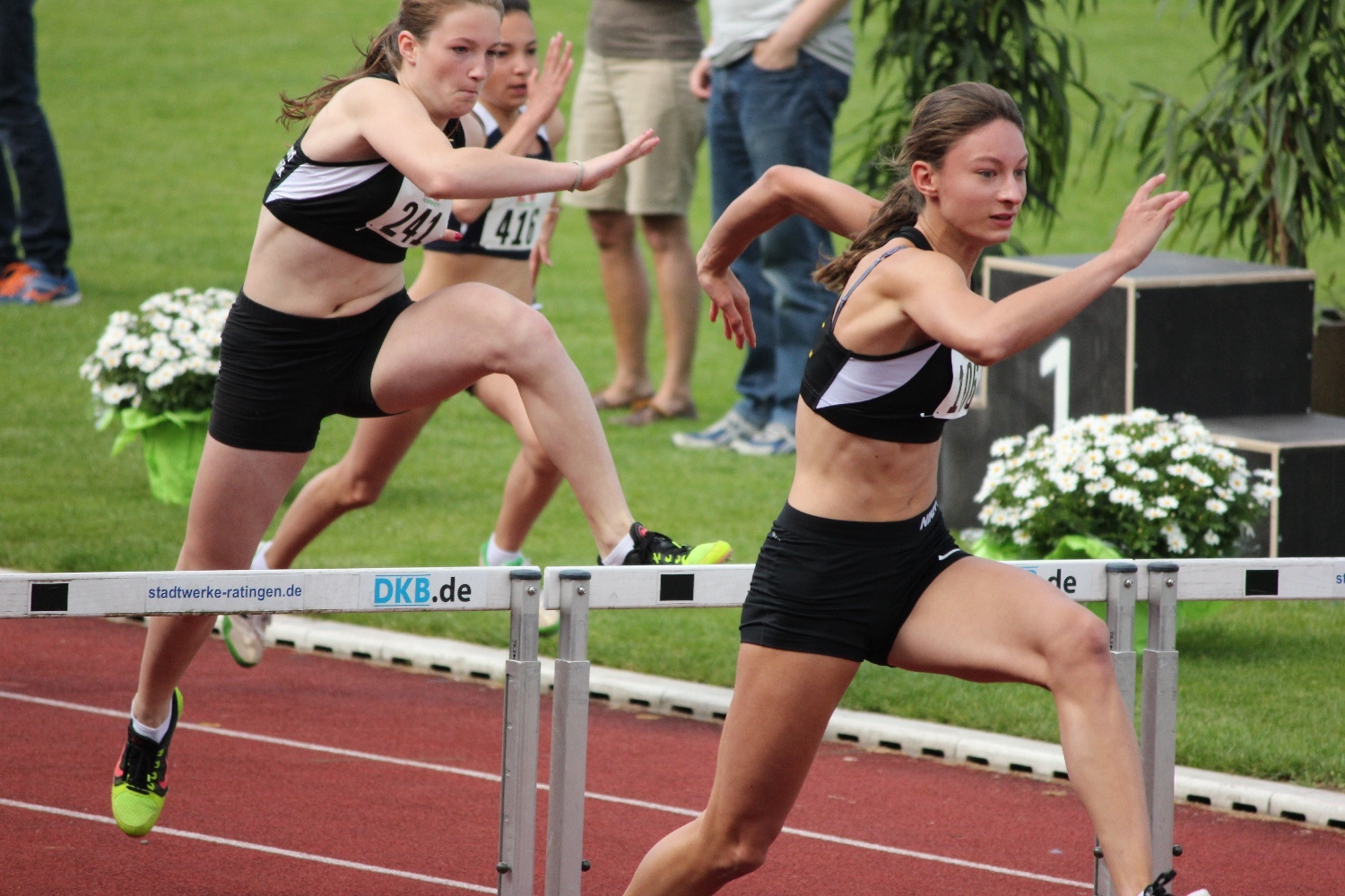 Zwei Athletinnen &uuml;berwinden Hindernisse beim H&uuml;rdenlauf auf einer roten Laufbahn. Zuschauer im Hintergrund.