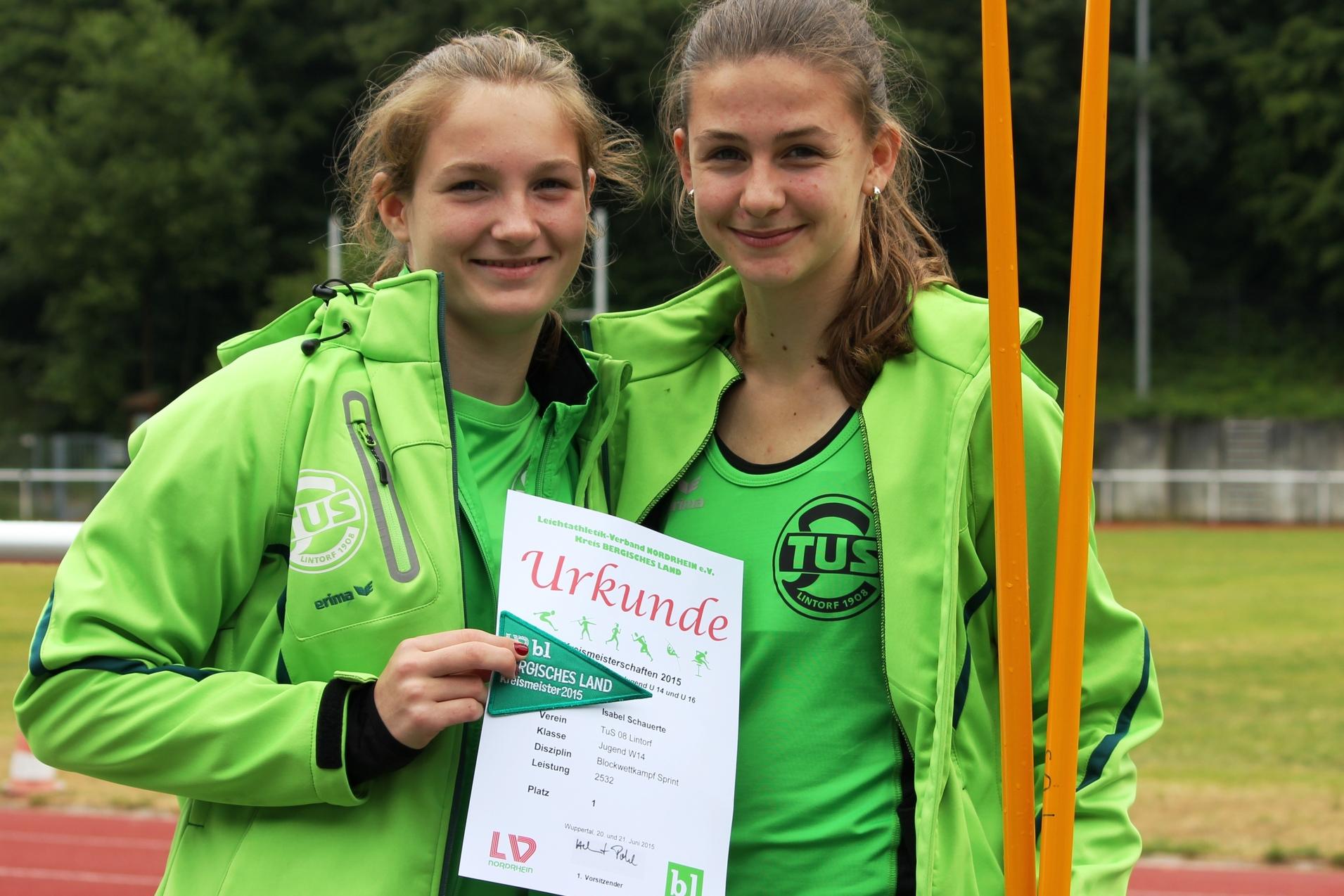 Zwei Jugendliche in grünen Sportjacken halten stolz eine Urkunde auf einem Sportplatz.