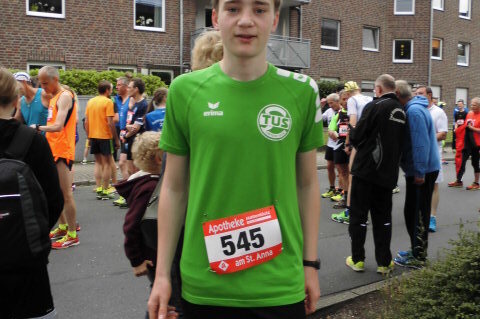 Junger Mann in gr&uuml;nem Sporttrikot mit Startnummer 545 steht in der Menge vor einem Wettkampf. Hintergrund mit L&auml;ufern und Geb&auml;uden.