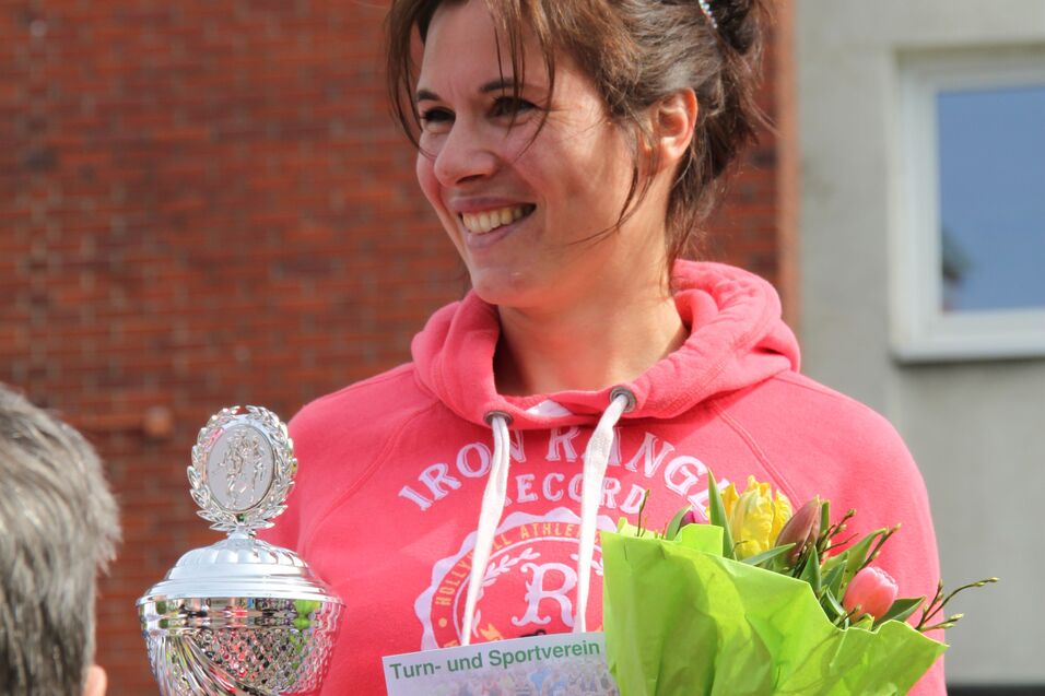 Frau mit Pokal und Blumenstrau&szlig;, l&auml;chelt, w&auml;hrend sie eine Urkunde in der anderen Hand h&auml;lt. Hintergrund aus rotem Ziegelstein.