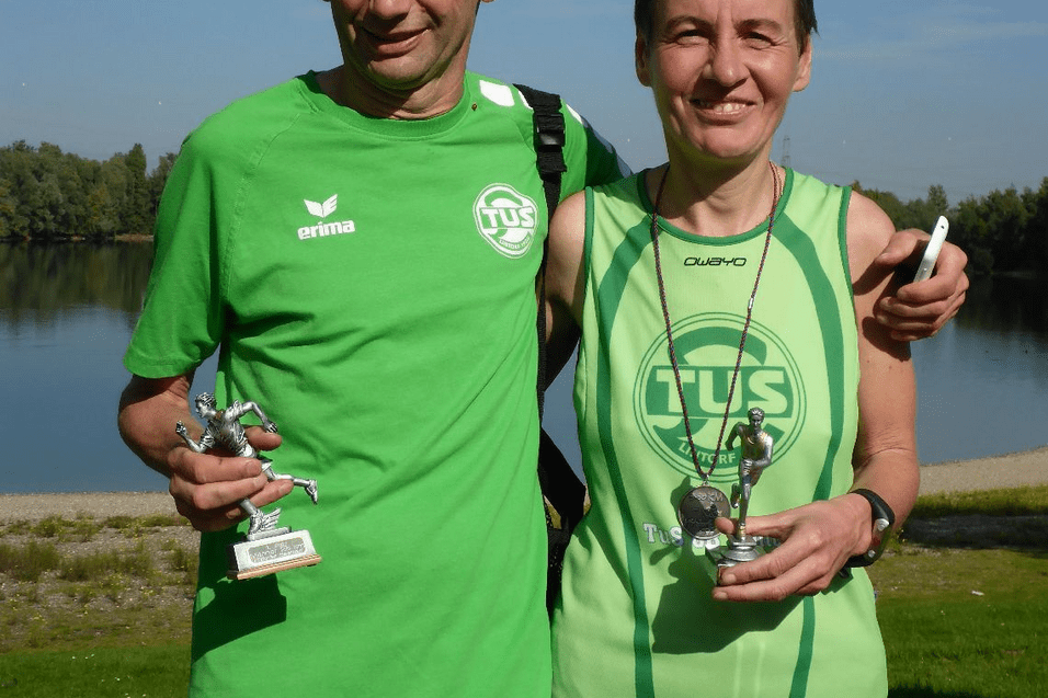 Zwei L&auml;ufer in Sportbekleidung halten Pokale vor einem Gew&auml;sser, umgeben von B&auml;umen.