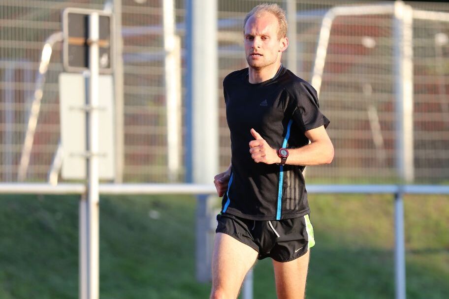 Mann in Sportbekleidung joggt auf einer roten Laufbahn, umgeben von einem gr&uuml;nen Landschaftshintergrund.