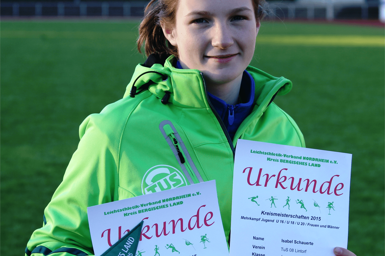 Junge Athletin in grüner Jacke hält Urkunden der Kreismeisterschaften 2015 auf einem Sportplatz.