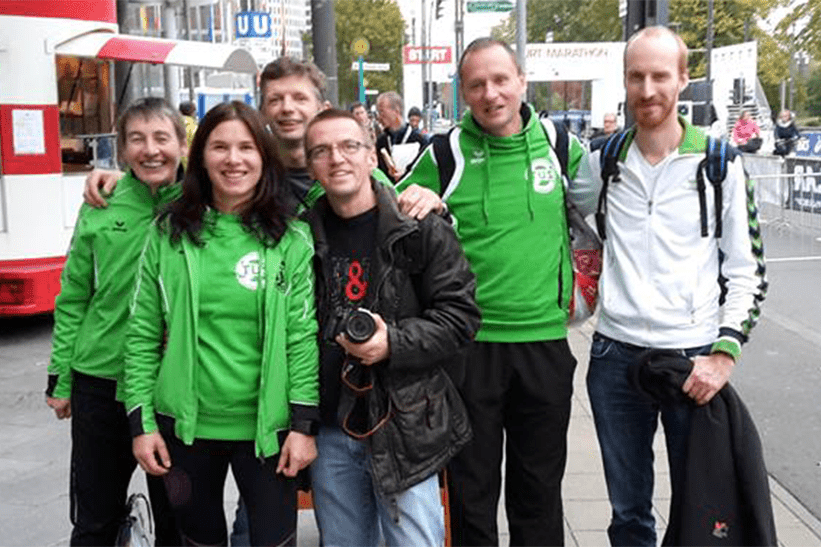 Gruppe von f&uuml;nf Personen in gr&uuml;nen Jacken l&auml;chelt vor einer urbanen Kulisse, eine Person h&auml;lt eine Kamera.