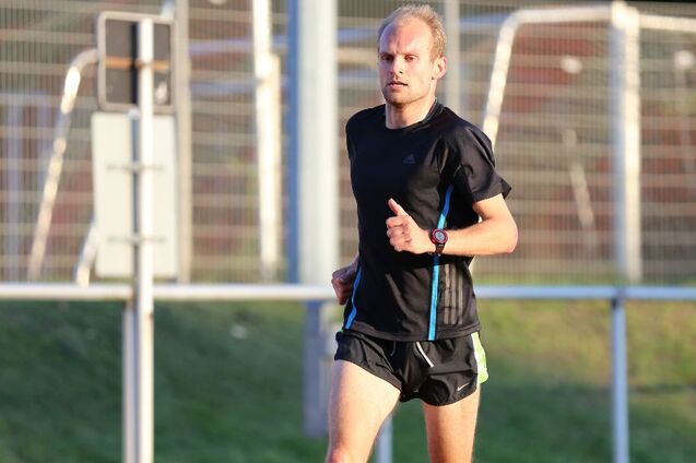 Mann in Sportbekleidung joggt auf einer roten Laufbahn, umgeben von einem gr&uuml;nen Landschaftshintergrund.