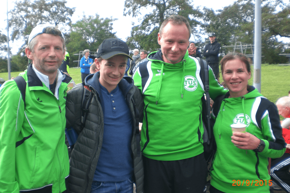 Vier Personen stehen auf einem Sportgel&auml;nde, alle tragen gr&uuml;ne Sportjacken. Im Hintergrund sind Zuschauer zu sehen.