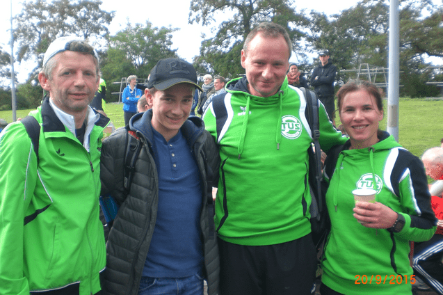 Vier Personen stehen auf einem Sportgel&auml;nde, alle tragen gr&uuml;ne Sportjacken. Im Hintergrund sind Zuschauer zu sehen.