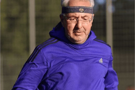 Mann in lila Sportswear und Stirnband l&auml;uft auf einem Sportplatz, im Hintergrund ist ein Zaun sichtbar.