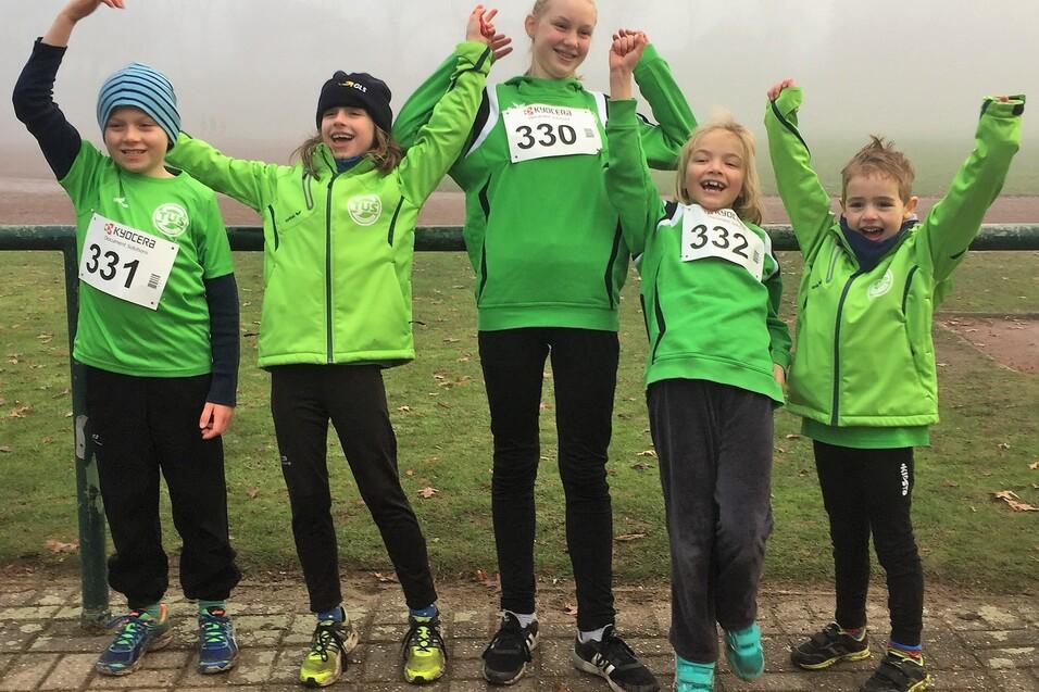 F&uuml;nf Kinder in gr&uuml;nen Sportjacken mit Startnummern l&auml;cheln und heben die Arme vor einem nebligen Sportplatz.