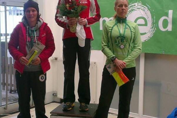 Drei Frauen stehen auf einem Siegertreppchen; die mittlere hält einen Blumenstrauß und trägt eine Medaille.