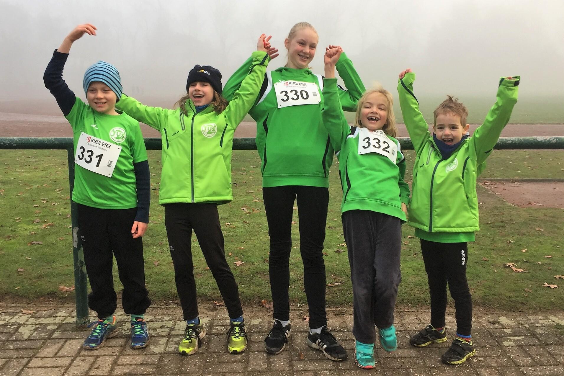 F&uuml;nf Kinder in gr&uuml;nen Sportjacken mit Startnummern l&auml;cheln und heben die Arme vor einem nebligen Sportplatz.