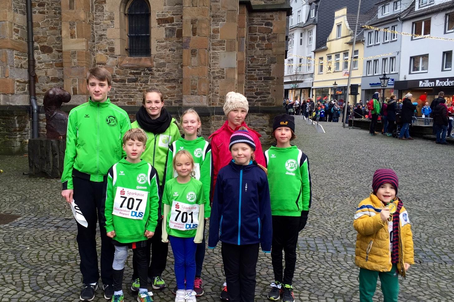 Gruppe von sieben Kindern in Sportkleidung mit Startnummern vor historischen Gebäuden auf einem weichen Pflaster.