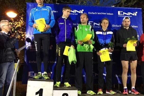 F&uuml;nf L&auml;ufer stehen auf dem Siegertreppchen, halten Urkunden und Lorbeerkr&auml;nze in der Hand. Hintergrund mit Logo und Beleuchtung.