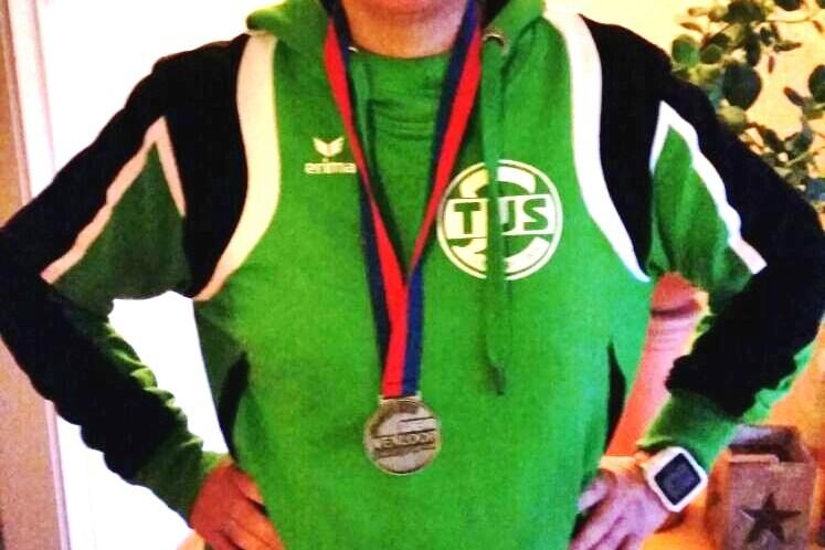 Frau in gr&uuml;ner Trainingsjacke mit Medaille um den Hals posiert in einem Raum mit Holzfu&szlig;boden und Pflanzen im Hintergrund.