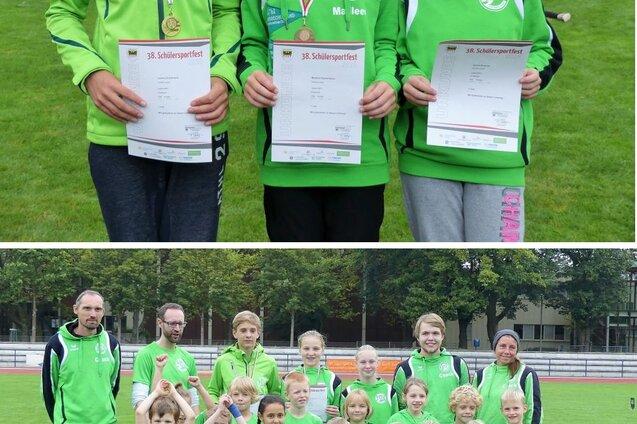 Drei Kinder mit Medaillen und Urkunden stehen auf einer Wiese; im Hintergrund eine Gruppe junger Sportler und Trainer.