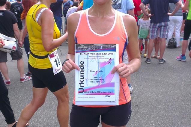 Frau mit Medaille und Urkunde steht vor einer Menschenmenge nach Volkslauf, umgeben von B&auml;umen und Verkaufsst&auml;nden.