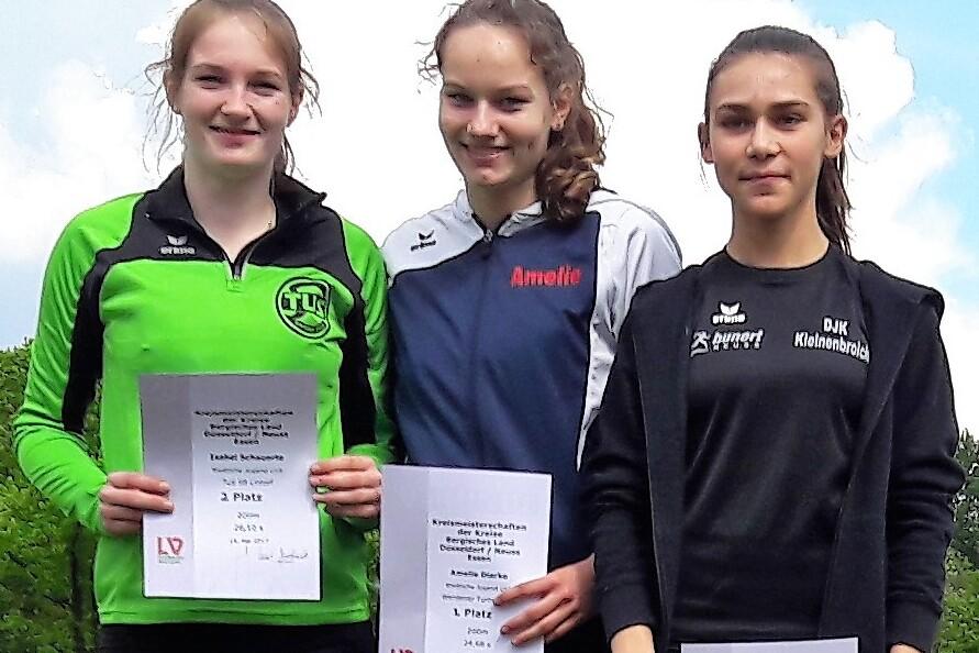 Drei junge Sportlerinnen stehen l&auml;chelnd mit Urkunden in der Hand vor einer gr&uuml;nen Landschaft.