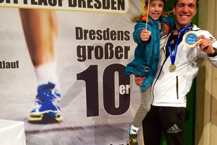 Mann mit Medaille h&auml;lt l&auml;chelnd ein Kind auf dem Arm vor einem Banner des Citylaufs Dresden.