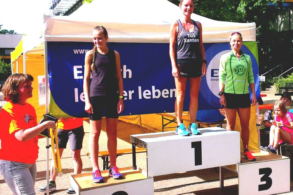 Drei Frauen auf dem Siegerpodest eines Wettkampfs, mit Platz 1 in der Mitte und Zuschauern im Hintergrund.