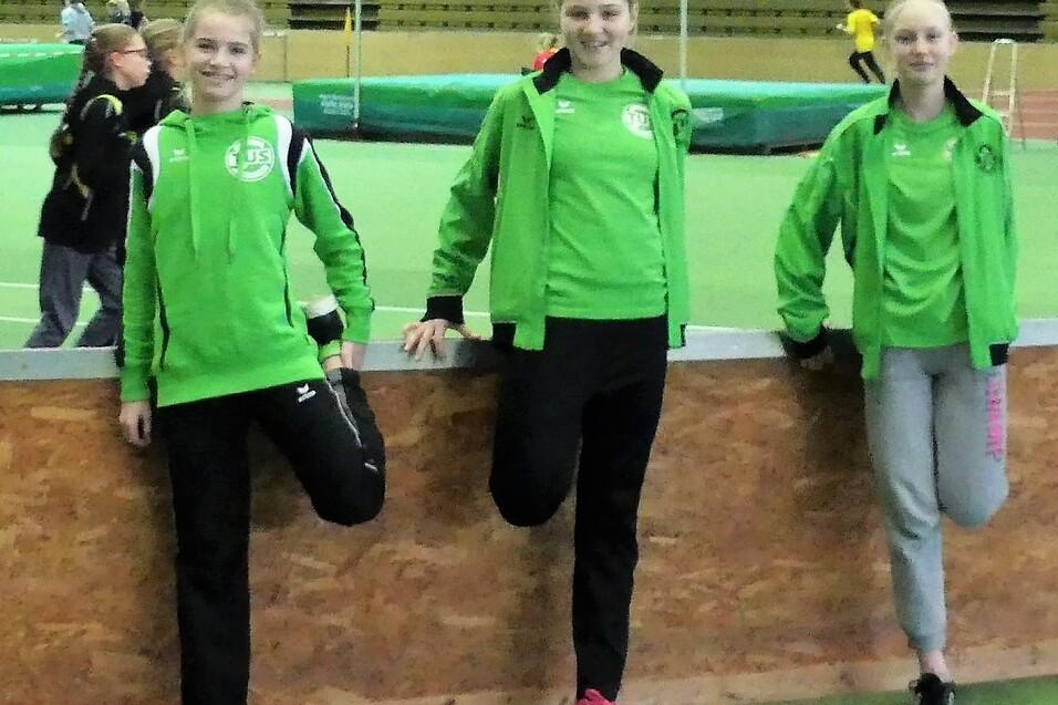 Drei sportliche M&auml;dchen in gr&uuml;ner Sportkleidung lehnen an einer Holzbarriere in einer Sporthalle.