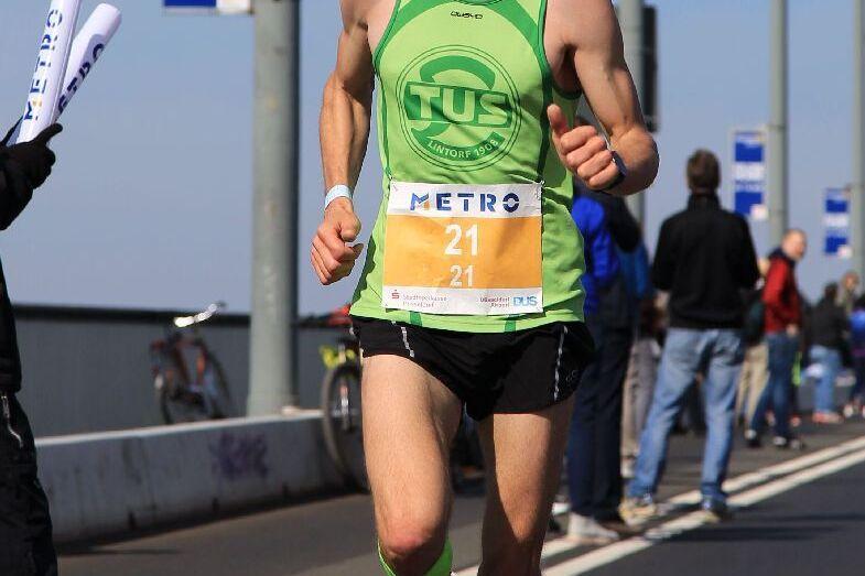 L&auml;ufer in gr&uuml;ner Sportbekleidung mit Startnummer 21 sprintet auf einer asphaltierte Stra&szlig;e, Zuschauer im Hintergrund.